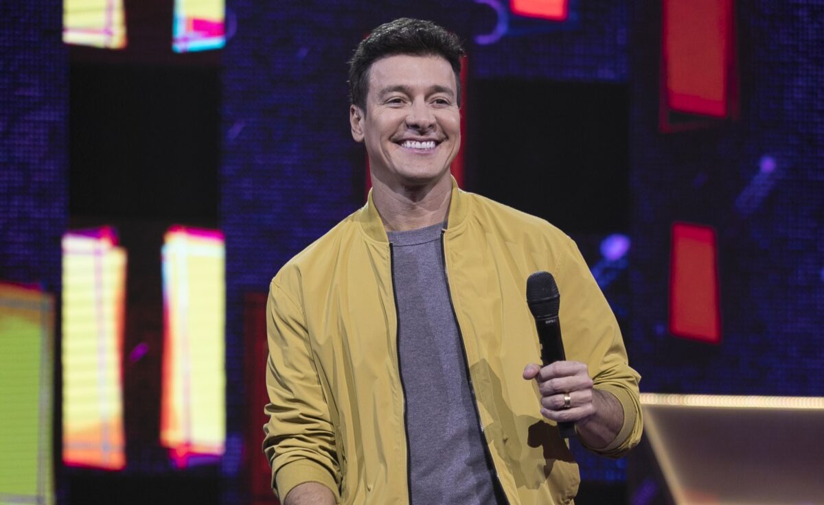 Rodrigo Faro segurando um microfone e sorridente, nos estúdios do Canta Comigo Teen