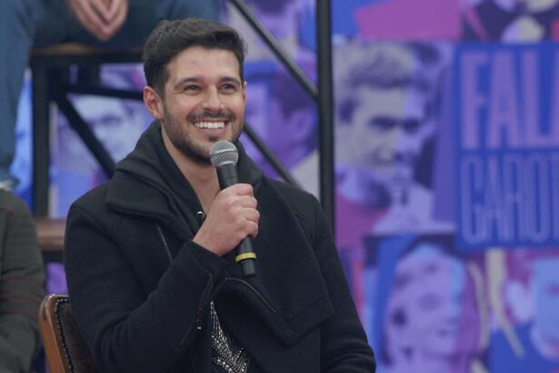 Rodrigo Mussi de jaqueta jeans e camisa preta, sentado, sorrindo, segurando o microfone com a mão direita durante entrevista no Altas Horas, da TV Globo