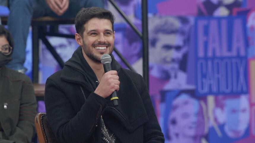 Rodrigo Mussi de jaqueta jeans e camisa preta, sentado, sorrindo, segurando o microfone com a mão direita durante entrevista no Altas Horas, da TV Globo