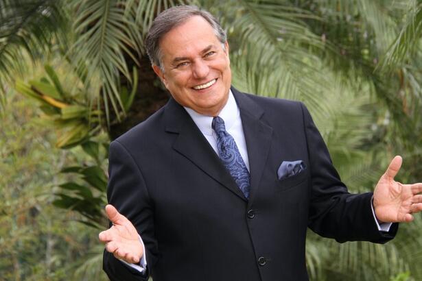 Ronnie Von de palitó preto e gravata e lenço em tons de ciano, sorrindo, com os braços abertos num espaço cercado por plantas durante ensaio fotográfico