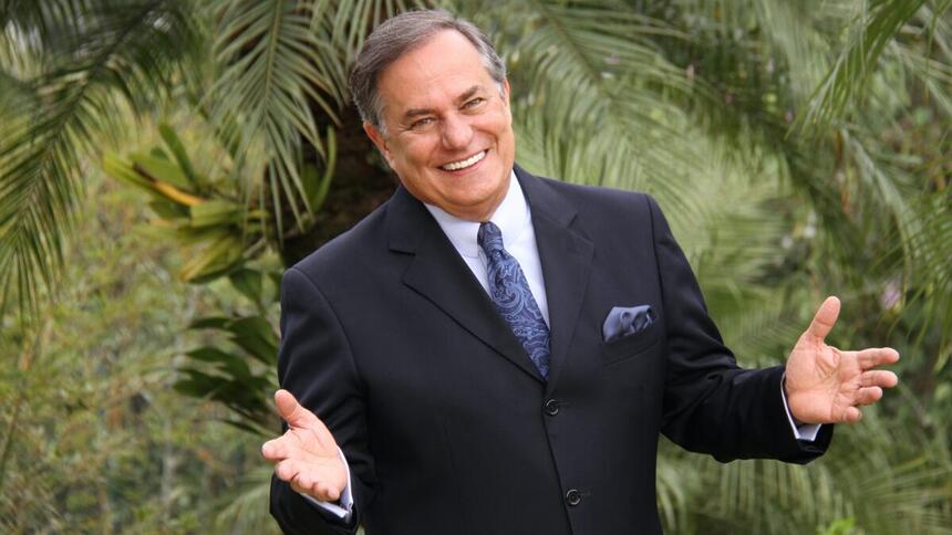 Ronnie Von de palitó preto e gravata e lenço em tons de ciano, sorrindo, com os braços abertos num espaço cercado por plantas durante ensaio fotográfico