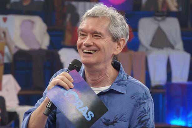 Serginho Groisman sorrindo segurando um microfone preto com a mão direita, usando camisa jeans azul por fora e uma tshirt branca por dentro, segurando a ficha do programa Altas Horas, da TV Globo