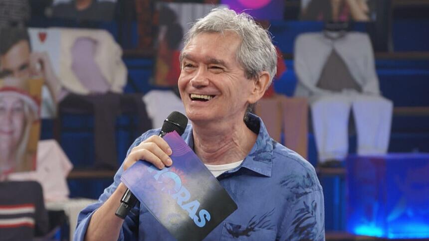 Serginho Groisman sorrindo segurando um microfone preto com a mão direita, usando camisa jeans azul por fora e uma tshirt branca por dentro, segurando a ficha do programa Altas Horas, da TV Globo