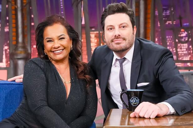 Solange Couto sorrindo, com as mãos juntas no joelho, usando macaquito preto brilhoso e um colar de brilhantes ao lado de Danilo gentili durante gravação do The Noite, do SBT