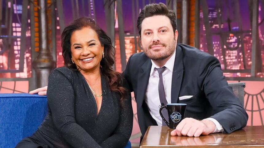 Solange Couto sorrindo, com as mãos juntas no joelho, usando macaquito preto brilhoso e um colar de brilhantes ao lado de Danilo gentili durante gravação do The Noite, do SBT