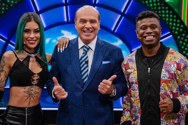 Tati Zaqui sorrindo, com look em decote preto, com a mão no ombro de Marcelo; Edilson Capetinha com blazer em estampa colorida e camisa preta, colar de prata, sorrindo; Marcelo de Carvalho no centro de blazer e gravata azul e camisa social branca fazendo um legal com as duas mãos durante gravação de O Céu é o Limite, da RedeTV!