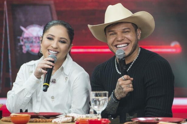 Mari Fernandez de jaqueta branca, microfone preto na mão direita, sorrindo, sentada ao lado de Tierry; Tierry de camisa manga longa preta, colar e chapéu de cowboy sorrindo durante gravação do Churrascão do Faustão, da Band