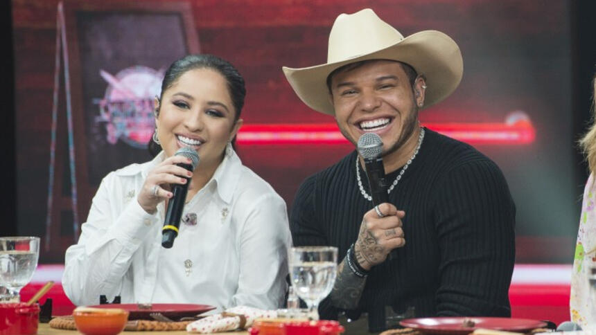 Mari Fernandez de jaqueta branca, microfone preto na mão direita, sorrindo, sentada ao lado de Tierry; Tierry de camisa manga longa preta, colar e chapéu de cowboy sorrindo durante gravação do Churrascão do Faustão, da Band