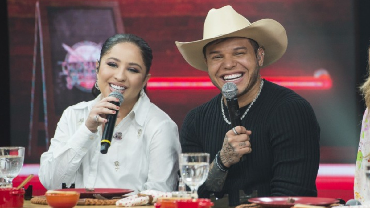 Mari Fernandez de jaqueta branca, microfone preto na mão direita, sorrindo, sentada ao lado de Tierry; Tierry de camisa manga longa preta, colar e chapéu de cowboy sorrindo durante gravação do Churrascão do Faustão, da Band