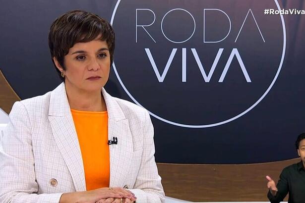 Vera Magalhães de blazer branco, camisa cinza, microfone preto grampeado no blazer, com semblante sério, sentada num banco durante gravação do Roda Viva, da TV Cultura