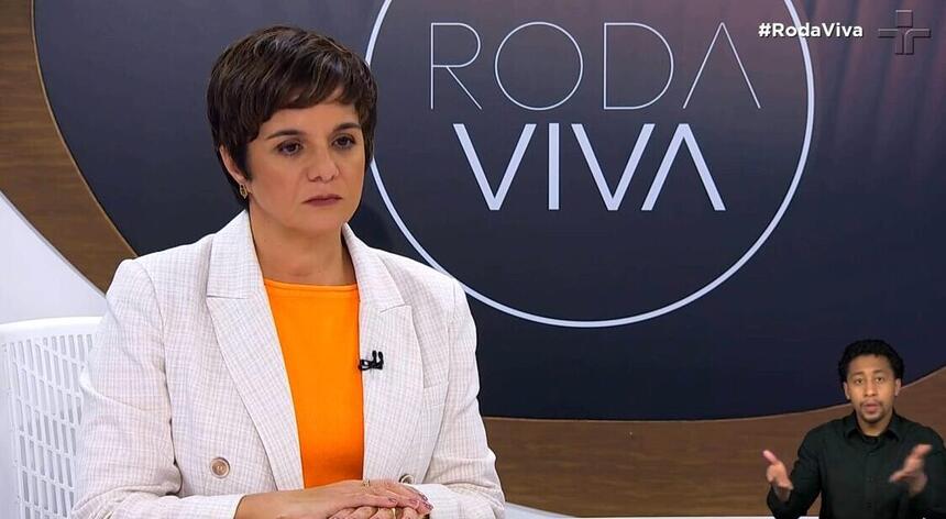 Vera Magalhães de blazer branco, camisa cinza, microfone preto grampeado no blazer, com semblante sério, sentada num banco durante gravação do Roda Viva, da TV Cultura