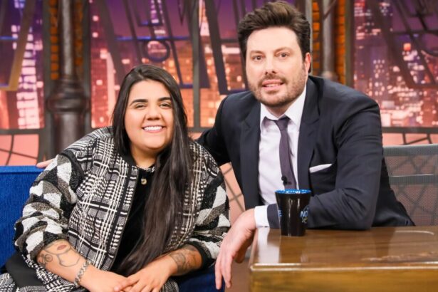 Yasmin Santos sorrindo, de camisa preta, sentada num sofá azul ao lado de Danilo Gentili durante gravação do The Noite, do SBT