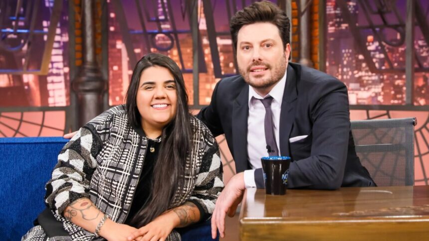 Yasmin Santos sorrindo, de camisa preta, sentada num sofá azul ao lado de Danilo Gentili durante gravação do The Noite, do SBT