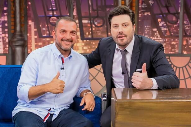 Zé Elias de camisa social azul clara e calça jeans, sorrindo, sentando no sofá azul, fazendo um legal com a mão direita; Danilo Gentili sorrindo, de palitó gravata e calça preta, sentado, fazendo um legal com a mão esquerda, na sua bancada durante gravação do The Noite, do SBT