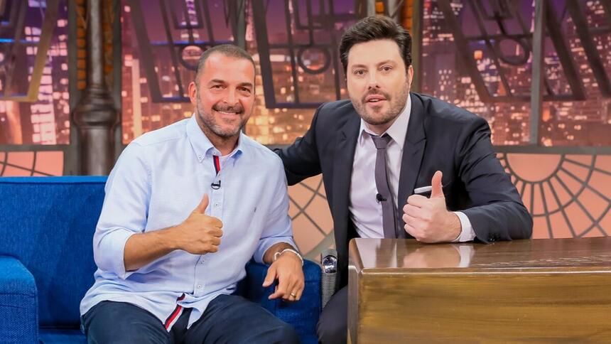 Zé Elias de camisa social azul clara e calça jeans, sorrindo, sentando no sofá azul, fazendo um legal com a mão direita; Danilo Gentili sorrindo, de palitó gravata e calça preta, sentado, fazendo um legal com a mão esquerda, na sua bancada durante gravação do The Noite, do SBT