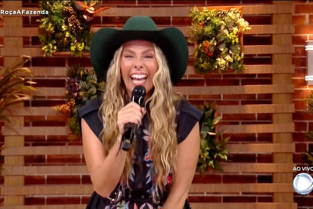 Adriane Galisteu, com chapéu na cabeça, microfone na mão e sorriso aberto
