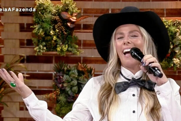 Adriane Galisteu sorrindo, de chapéu de cowboy preto, laço e camisa social, segurando o microfone preto