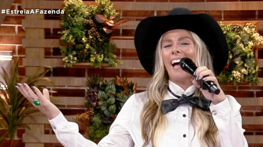 Adriane Galisteu sorrindo, de chapéu de cowboy preto, laço e camisa social, segurando o microfone preto