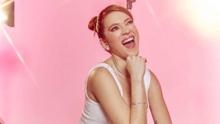 Ana Clara de cabelo presa, sorrindo, mão no queixo, camisa regata, num estúdio rosa