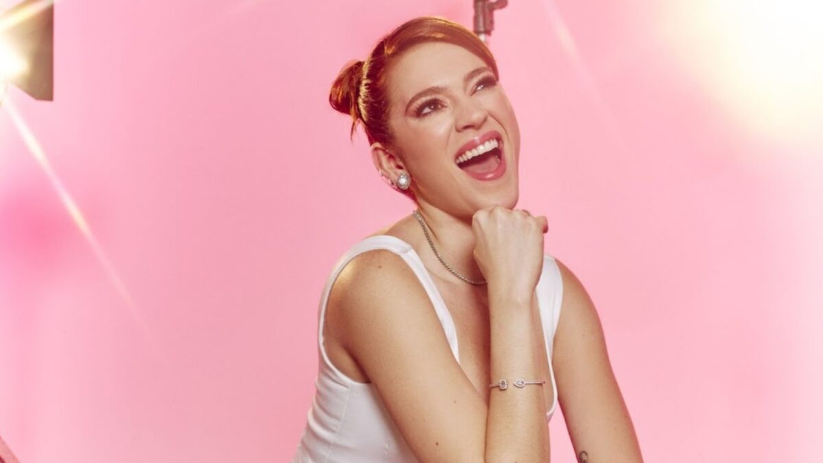 Ana Clara de cabelo presa, sorrindo, mão no queixo, camisa regata, num estúdio rosa