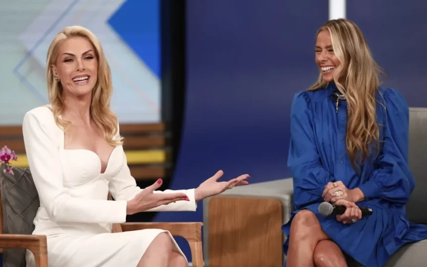 Ana Hickmann de vestido branco, com plaquinhas no banco, sorrindo, de cabelo solto, ao lado de Adriane Galisteu; Adriane Galisteu de vestido azul, cabelo solto, sorrindo durante entrevista em coletiva de A Fazenda no Hoje em Dia, da Record TV