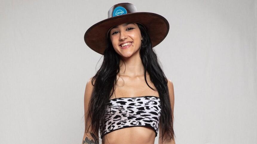 Bia Miranda sorrindo, de cabelo solto, top em estampa de vaca , com chapéu de fazendeiro durante ensaio fotográfico para A Fazenda 14