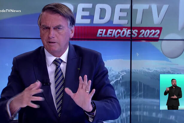 Bolsonaro de palitó, preto, gravata listrada preta a branca, camisa social branca, gesticulando com as mãos, sentado durante entrevista na RedeTV!