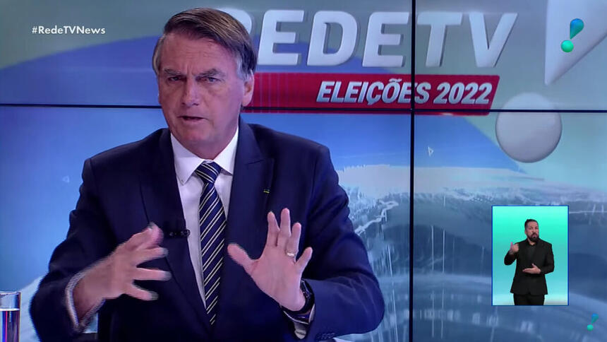 Bolsonaro de palitó, preto, gravata listrada preta a branca, camisa social branca, gesticulando com as mãos, sentado durante entrevista na RedeTV!