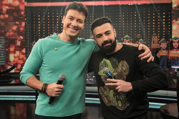 Rodrigo Faro de camisa verde, sorrindo, segurando o microfone; Bruno Tálamo de camisa preta estampada durante gravação do Hora do Faro, da Record TV