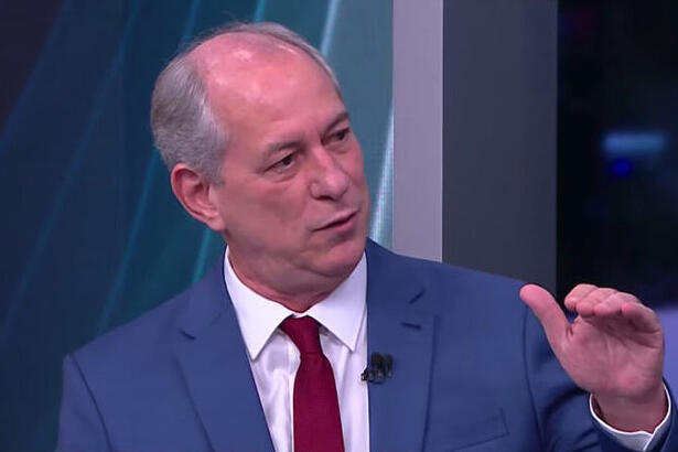 Ciro Gomes de palitó azul, gravata vermelha e camisa social branca, sentado, com a mão pra cima durante entrevista na CNN Brasil