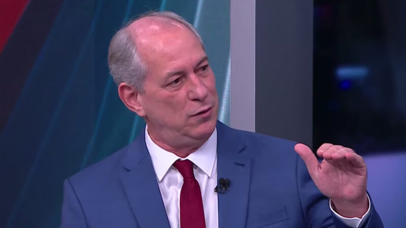 Ciro Gomes de palitó azul, gravata vermelha e camisa social branca, sentado, com a mão pra cima durante entrevista na CNN Brasil