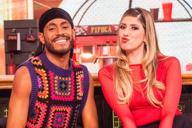 ícaro Silva de bandana preta e camisa de crochê estampada, sorrindo, ao lado de Dani Calabresa, de camisa vermelha e saia preta, batom vermelho e cabelo solto durante gravação do Domingo com Huck, da TV Globo