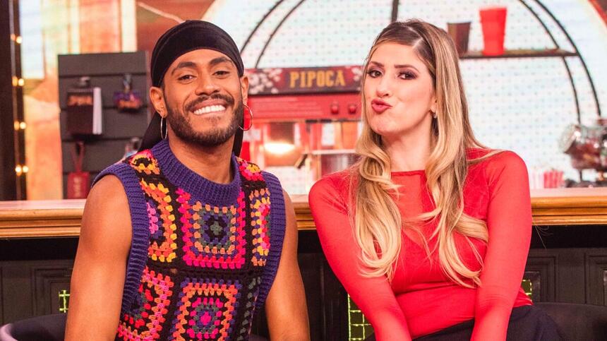 ícaro Silva de bandana preta e camisa de crochê estampada, sorrindo, ao lado de Dani Calabresa, de camisa vermelha e saia preta, batom vermelho e cabelo solto durante gravação do Domingo com Huck, da TV Globo