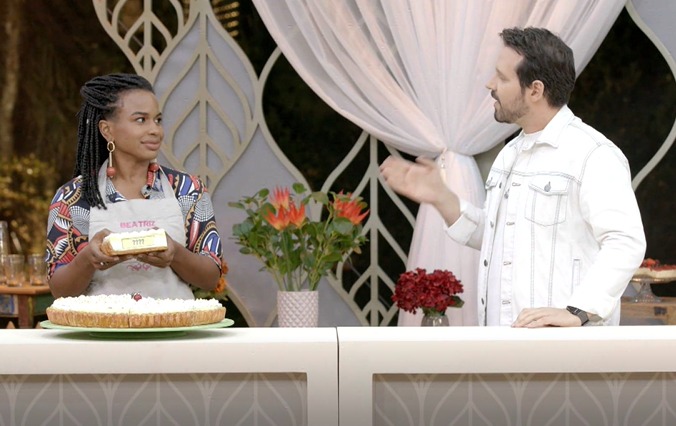 Beatriz de avental segurando um bolo com a mão enquanto conversa com Dony De Nuccio durante gravação do Bake Off Brasil