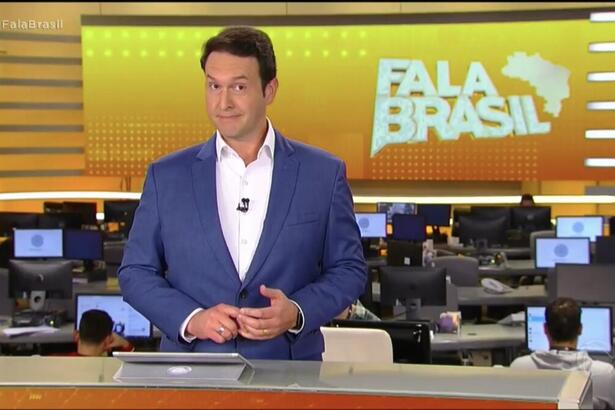 Eduardo Ribeiro, de terno sem gravata, atrás da bancada nos estúdios do Fala Brasil, com a redação de jornalismo ao fundo e o telão com o logo do telejornal