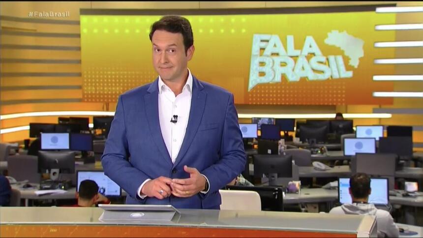 Eduardo Ribeiro, de terno sem gravata, atrás da bancada nos estúdios do Fala Brasil, com a redação de jornalismo ao fundo e o telão com o logo do telejornal