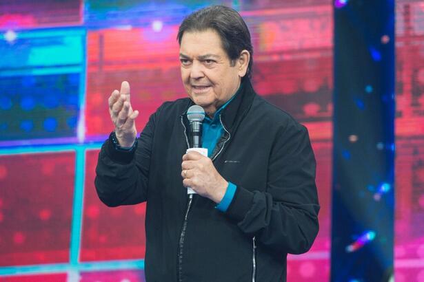 Faustão de colete preto, camisa azul, sorrindo, com microfone e ficha na mão esquerda durante gravação do seu programa na Band