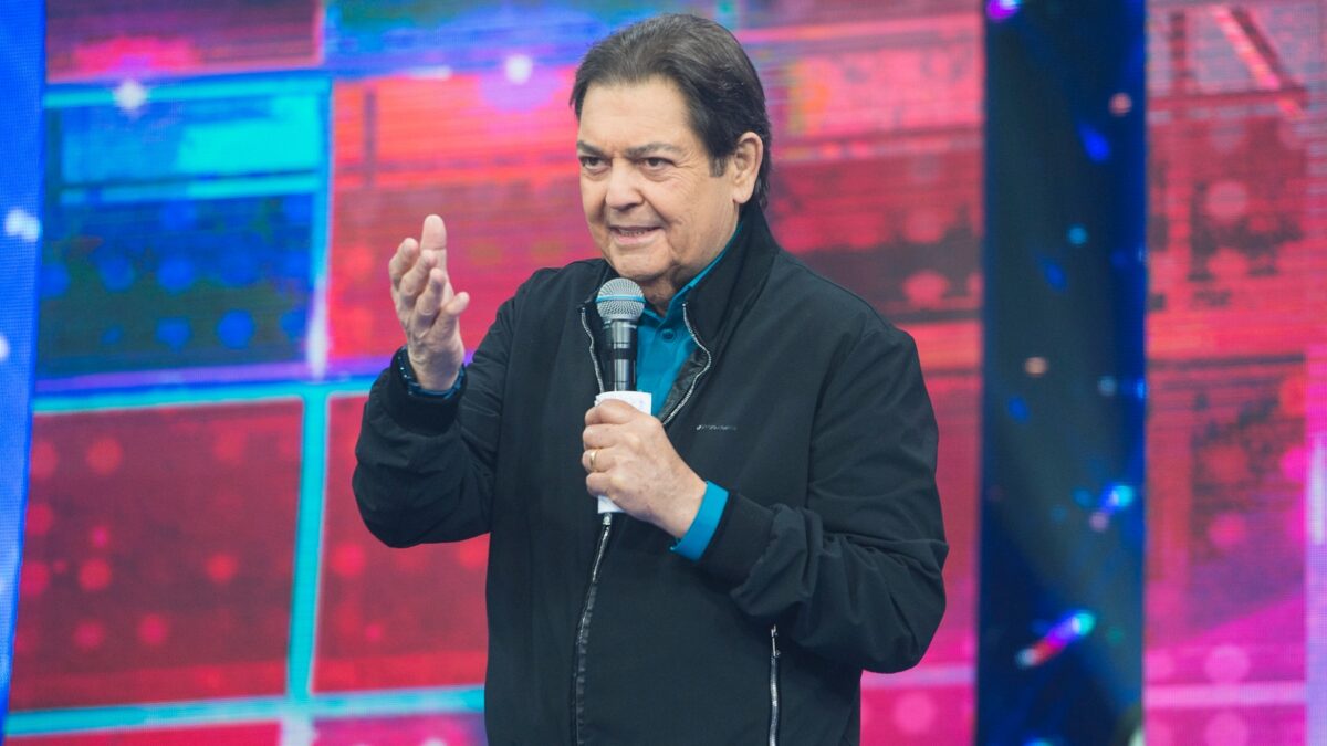 Faustão de colete preto, camisa azul, sorrindo, com microfone e ficha na mão esquerda durante gravação do seu programa na Band