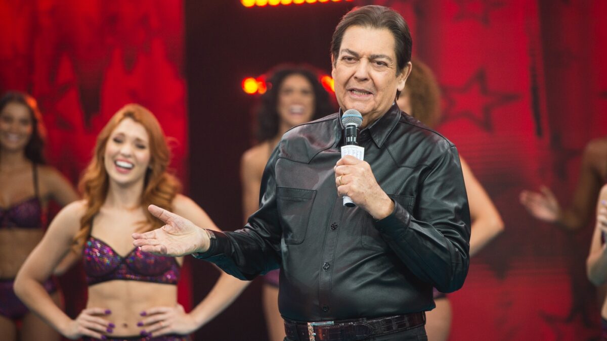 Faustão de jaqueta de couro e calça preta com os braços abertos, segurando um microfone e fichas durante apresentação do seu programa