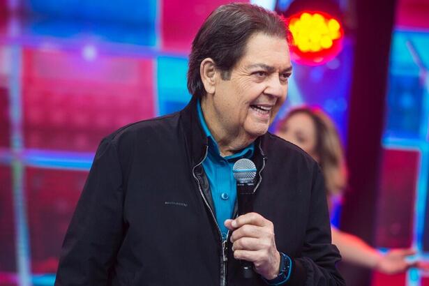 Fausto Silva de jaqueta jeans preta, camisa social azul, com microfone na mão esquerda durante gravação do Faustão na Band, da Band