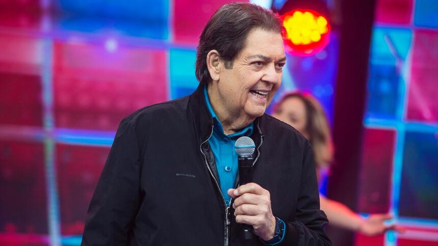 Fausto Silva de jaqueta jeans preta, camisa social azul, com microfone na mão esquerda durante gravação do Faustão na Band, da Band