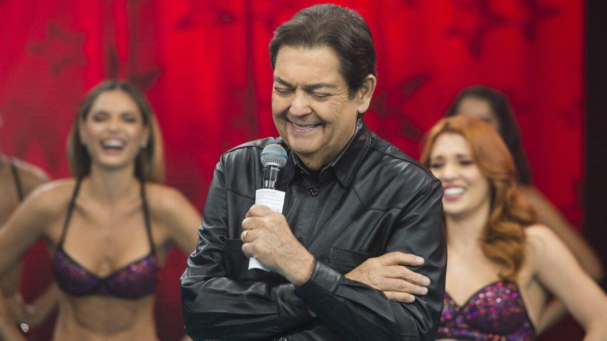 Faustão de jaqueta de couro preta, segurando um microfone com a mão esquerda, de braços cruzados, duranta gravação do seu programa na Band