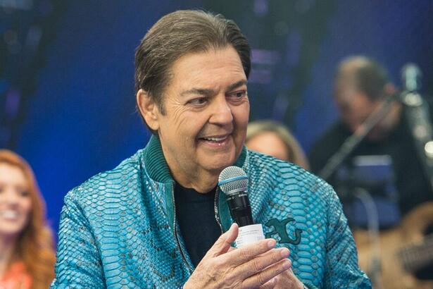 Fausto Silva de camisa mangalonga azul e camisa preta, com o microfone e ficha na mão, sorrindo durante gravação do Faustão na Band