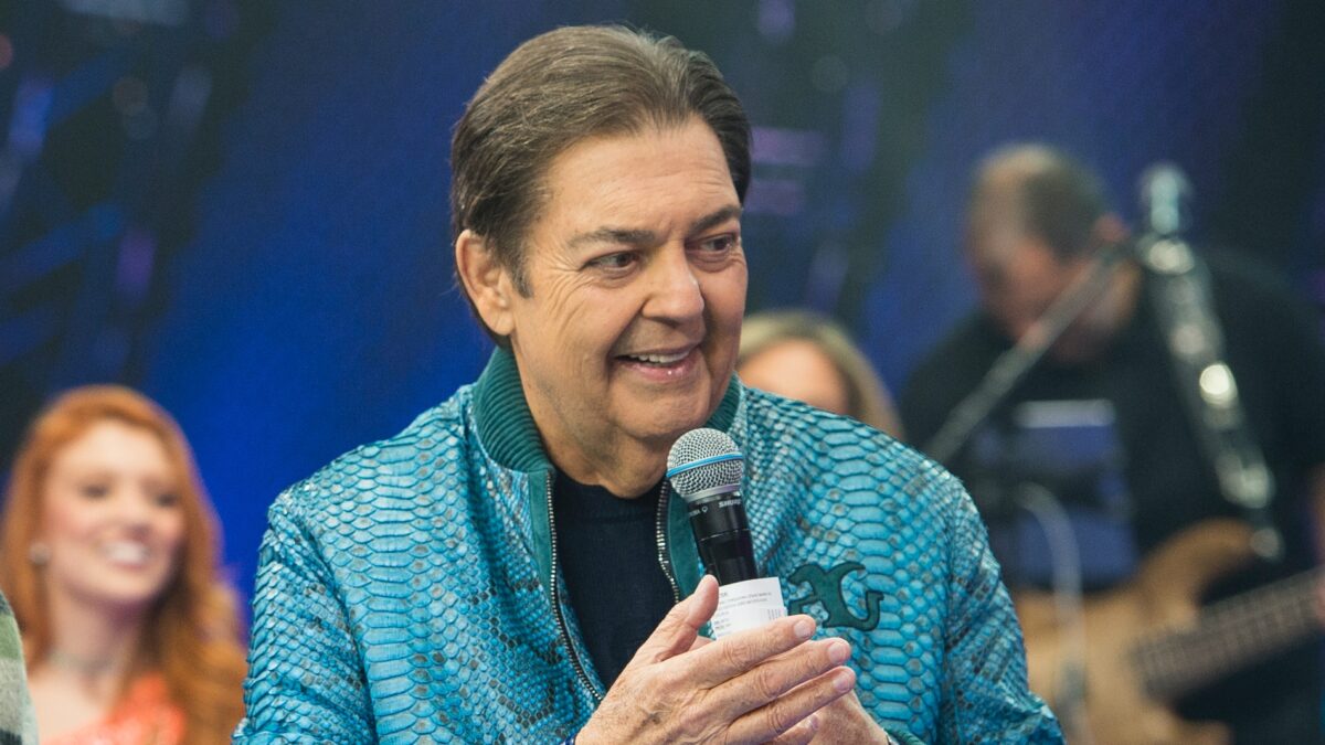 Fausto Silva de camisa mangalonga azul e camisa preta, com o microfone e ficha na mão, sorrindo durante gravação do Faustão na Band
