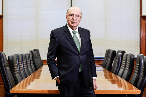 Henrique Meirelles de terno e gravata, se posionado em frente a uma mesa de reunião com várias cadeiras