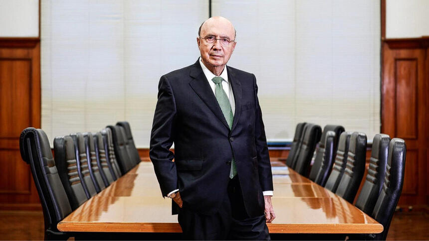 Henrique Meirelles de terno e gravata, se posionado em frente a uma mesa de reunião com várias cadeiras