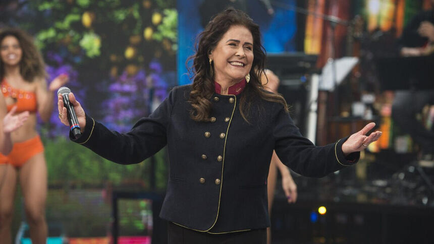 Joanna de camisa manga longa preta, com os braços abertos, segurando o microfone durante gravação do Faustão na Band