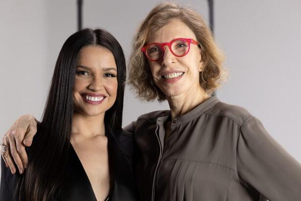 Marília Gabriela de camisa social cinza, sorrindo, de óculos de sol vermelho, cabelo solto ao lado de Juliette, de camisa preta, decote, sorrindo e cabelo solto durante gravação do programa