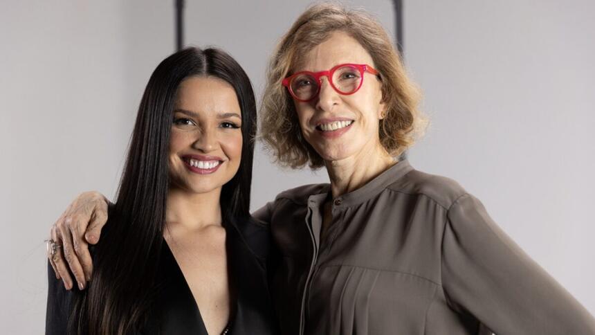 Marília Gabriela de camisa social cinza, sorrindo, de óculos de sol vermelho, cabelo solto ao lado de Juliette, de camisa preta, decote, sorrindo e cabelo solto durante gravação do programa