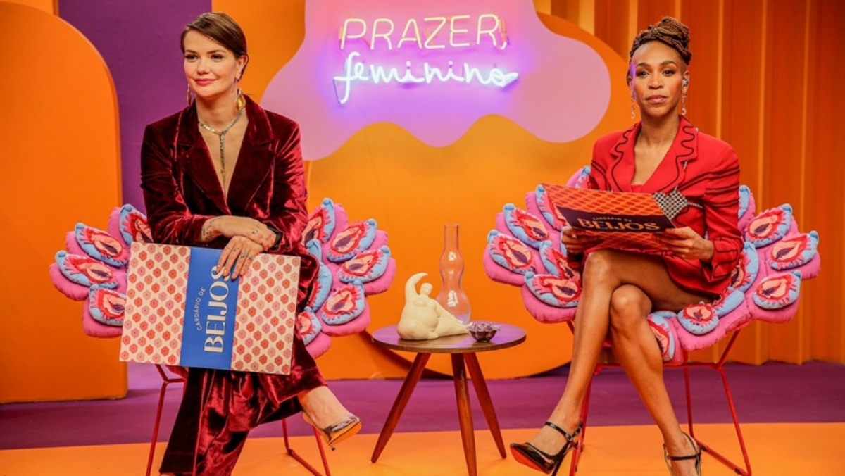 Marcela McGowan de blazer e calça em tons de magenta saturado, segurando placa do programa, sentada num banco de madeira; Karol Conka de blazer e saia vermelha, sentada num banco de madeira durante gravação do Prazer, Feminino, do GNT